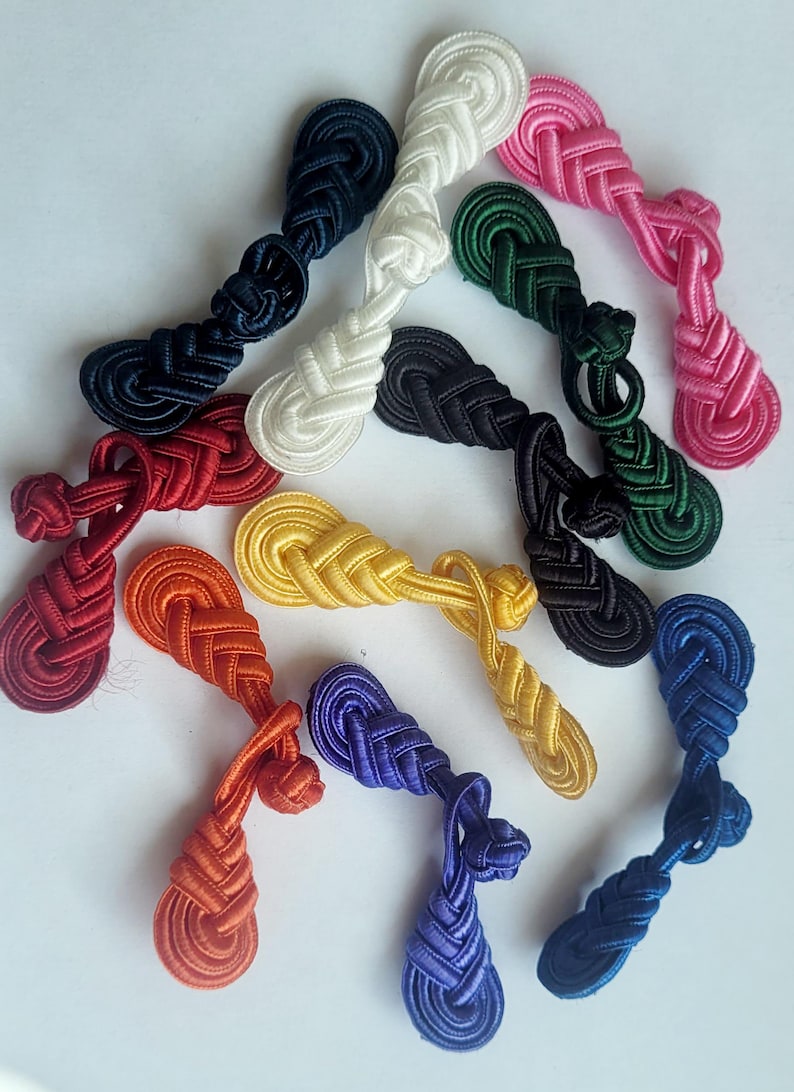 K&ouml;nnte beinhalten: Sortiment von 12 bunten, gekn&uuml;pften Stoff-Kn&ouml;pfen oder -Verschl&uuml;ssen. Die Kn&ouml;pfe sind in verschiedenen Farben, darunter Rot, Orange, Gelb, Gr&uuml;n, Blau, Wei&szlig; und Pink.