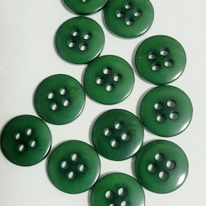 May include: A collection of eleven round, dark green buttons, each featuring a smooth surface and four centered holes for attachment. The buttons are arranged in a scattered pattern, displaying a consistent circular shape and uniform color.
