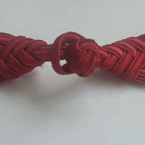 K&ouml;nnte beinhalten: Ein roter Stoffknopf mit geflochtenem Design. Der Knopf ist in der Mitte verknotet.