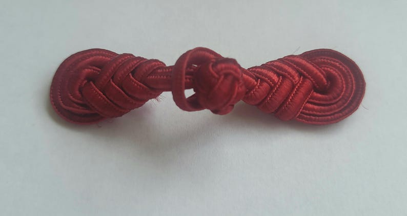 K&ouml;nnte beinhalten: Ein roter Stoffknopf mit geflochtenem Design. Der Knopf ist in der Mitte verknotet.