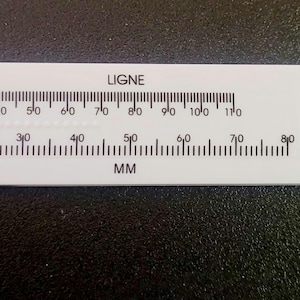 May include: A white plastic caliper with black measurement markings in millimeters. The caliper has a sliding scale and a button-like feature with four holes. The text "LIGNE" and "MM" are printed on the caliper.
