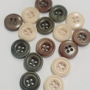 May include: A selection of round buttons in shades of brown, beige, and green. Each button has four holes for sewing. The buttons are displayed on a white surface, highlighting their different colours and textures.
