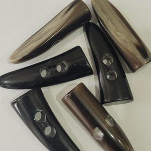Puede incluir: Una colección de botones de cuerno en varios tonos de marrón y negro. Los botones son alargados con un extremo redondeado y dos agujeros para coser. Algunos botones tienen un aspecto veteado.