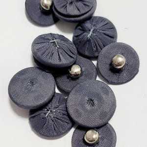 May include: A selection of dark grey fabric-covered buttons with silver metal centres. The buttons are circular and have a textured appearance, with a slightly raised centre. The buttons are arranged on a white surface.