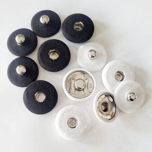 Puede incluir: Una colección de broches de presión cubiertos de tela negra y blanca con centros de metal plateado. Los broches son redondos y de diferentes tamaños, algunos muestran el mecanismo de cierre metálico. Los cierres están dispuestos sobre una superficie blanca.