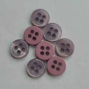 Peut inclure: Un ensemble de huit boutons ronds dans des tons de rose et de lavande. Chaque bouton comporte quatre trous pour la couture. Les boutons sont dispos&eacute;s sur une surface blanche, mettant en valeur leur couleur et leur design.