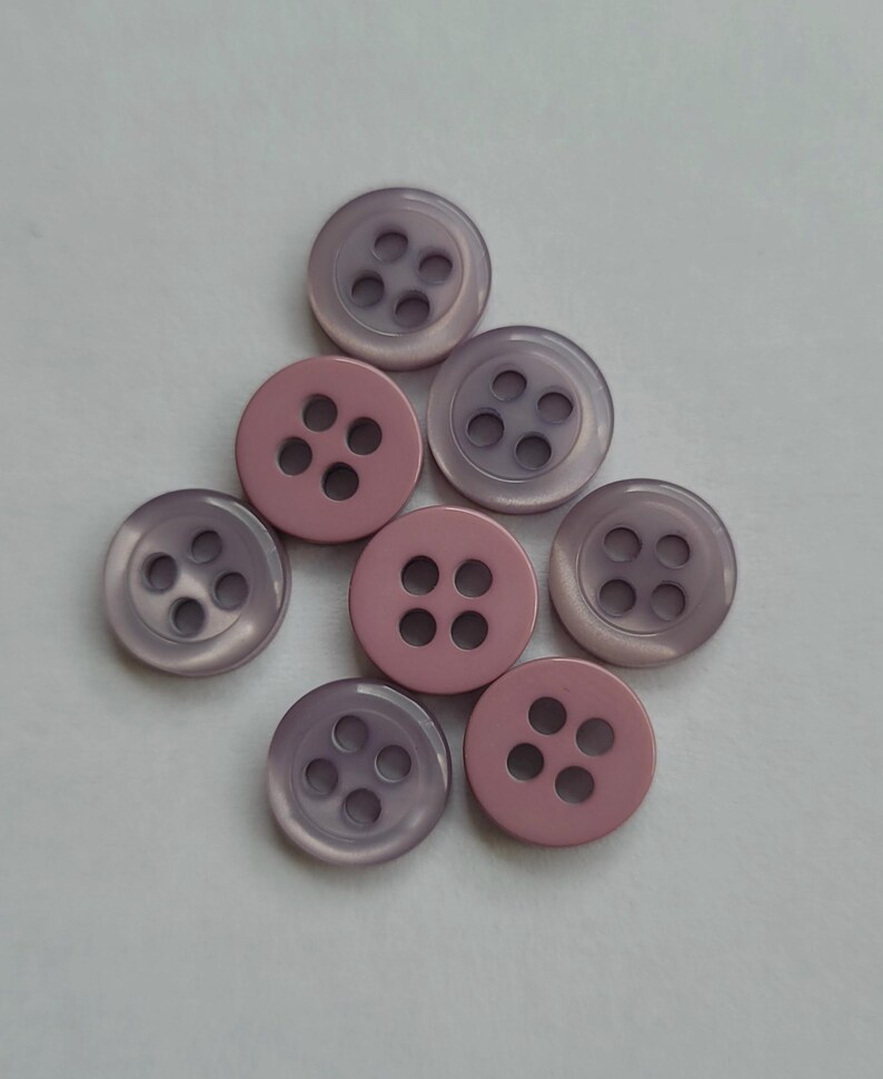 Peut inclure: Un ensemble de huit boutons ronds dans des tons de rose et de lavande. Chaque bouton comporte quatre trous pour la couture. Les boutons sont dispos&eacute;s sur une surface blanche, mettant en valeur leur couleur et leur design.