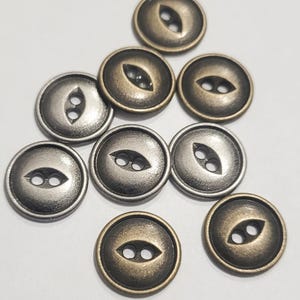 Peut inclure: Plusieurs boutons métalliques de couleurs bronze et argent. Chaque bouton est rond avec deux petits trous et une fente décorative en forme d'œil. Les boutons sont disposés sur une surface blanche.