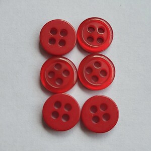 Peut inclure: Six boutons ronds rouges avec quatre trous chacun, dispos&eacute;s en deux rang&eacute;es de trois. Les boutons sont faits d'un mat&eacute;riau translucide, sur fond blanc.