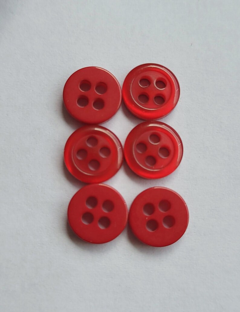 Peut inclure: Six boutons ronds rouges avec quatre trous chacun, dispos&eacute;s en deux rang&eacute;es de trois. Les boutons sont faits d'un mat&eacute;riau translucide, sur fond blanc.