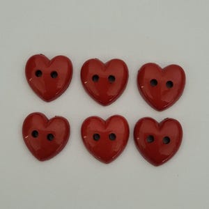 May include: Six red heart-shaped buttons with two holes each. The buttons are made of plastic and have a glossy finish.