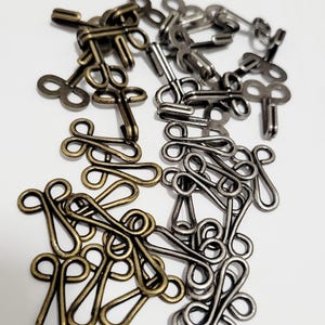 Op de afbeelding: Een verzameling metalen haak- en oogsluitingen in antiek messing en zilverkleurige tinten. De sluitingen zijn in verschillende maten en vormen, geschikt voor naai- en knutselprojecten. De haken hebben een lusvormig ontwerp.