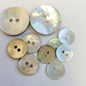 May include: A selection of iridescent, circular buttons in different sizes. Each button has two holes and shows a spectrum of colours, including white, cream, and touches of green and gold. These buttons are suitable for sewing and craft activities.