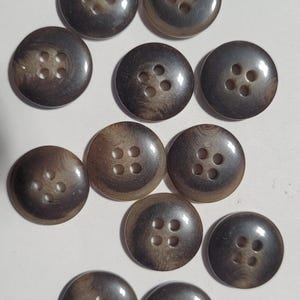 May include: A collection of eleven round, brown buttons with four holes each. The buttons have a marbled appearance with shades of brown and tan. They are arranged in a scattered pattern.