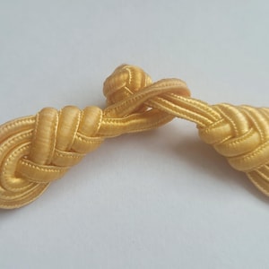 K&ouml;nnte beinhalten: Ein Paar goldfarbene Stoffkn&ouml;pfe, jeweils mit einem geflochtenen und geknoteten Design. Die Kn&ouml;pfe sind durch einen einzigen Faden Stoff verbunden.