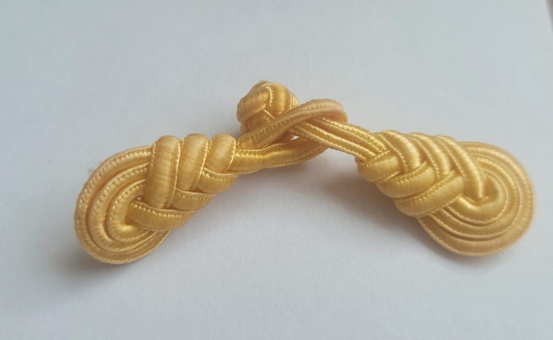 K&ouml;nnte beinhalten: Ein Paar goldfarbene Stoffkn&ouml;pfe, jeweils mit einem geflochtenen und geknoteten Design. Die Kn&ouml;pfe sind durch einen einzigen Faden Stoff verbunden.