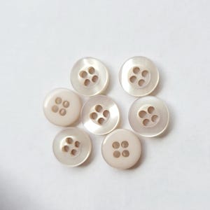 Peut inclure: Sept boutons ronds blanc cass&eacute;, chacun perc&eacute; de quatre trous, sont dispos&eacute;s sur un fond blanc. Les boutons ont un &eacute;clat nacr&eacute;, refl&eacute;tant la lumi&egrave;re. Les boutons sont de conception classique, adapt&eacute;s &agrave; divers projets de couture.