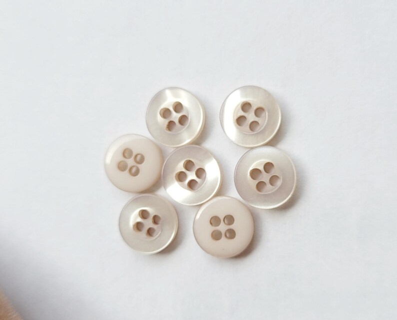Peut inclure: Sept boutons ronds blanc cass&eacute;, chacun perc&eacute; de quatre trous, sont dispos&eacute;s sur un fond blanc. Les boutons ont un &eacute;clat nacr&eacute;, refl&eacute;tant la lumi&egrave;re. Les boutons sont de conception classique, adapt&eacute;s &agrave; divers projets de couture.