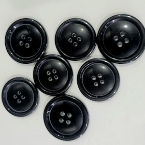 May include: Seven glossy black buttons of different sizes are displayed. Each button features four sewing holes and a subtly raised centre. The buttons are arranged on a white surface, highlighting their smooth, reflective surfaces.