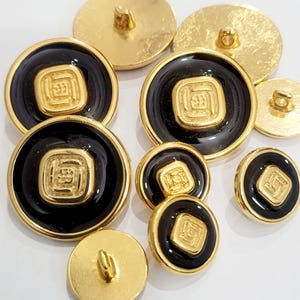 Peut inclure: Une collection de boutons ronds présentant un centre noir brillant avec un emblème carré doré en relief au milieu, entouré d'un bord doré. Certains boutons sont montrés de dos, révélant une attache à tige de couleur dorée.