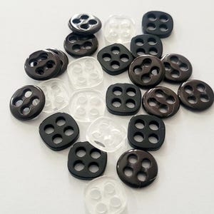 Op de afbeelding: Een verzameling kleine knopen met vier gaten van zwart en doorzichtig plastic. Het assortiment bevat zowel ronde als vierkante knopen met gladde, glanzende oppervlakken, verspreid gerangschikt op een effen witte ondergrond.