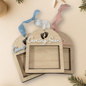 May include: Three wooden ornaments with a window frame and the text "Coming Soon" in white lettering. The ornaments are decorated with blue, pink, and white ribbons.