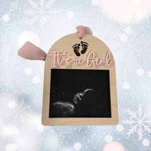 May include: Wooden sign with a pink ribbon, a picture frame, and the text "It's a Girl" in pink letters. The frame contains a black and white ultrasound image.