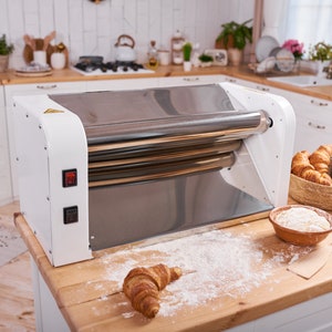 Peut inclure: Une machine à pâtes à rouler en acier inoxydable et blanche avec deux boutons rouges. La machine est sur un comptoir en bois avec un bol de farine, un racloir à pâte en bois et plusieurs croissants.