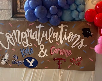 May include: A brown rectangular banner featuring the white hand-lettered text Congratulations, with a black graduation cap icon. It includes the names Kate and Caroline, the phrases go cougs and guns up, and logos for BYU and Texas Tech, accented by colorful sprinkles.