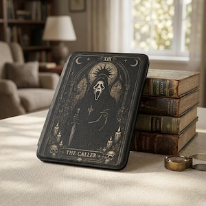 May include: Black case with a gothic design of a hooded figure, skulls, and candles. The text "XIII" and "THE CALLER" are displayed. The case is on a table next to a stack of old books.