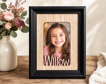 May include: A black framed photo with a light wood interior. The photo features a smiling girl with the name "Willow" cut out of the wood. A vase of flowers sits to the left of the frame.