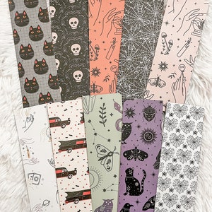 May include: A collection of decorative bookmarks with various spooky and Halloween-themed designs. The bookmarks feature illustrations of black cats, skulls, spiderwebs, hands, bats, and other mystical elements. The color palette includes black, gray, orange, and purple.