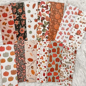 May include: A collection of rectangular paper bookmarks with various autumn-themed patterns. Designs include pumpkins, sunflowers, leaves, and acorns in shades of orange, brown, and cream. The bookmarks are arranged on a white, textured surface.