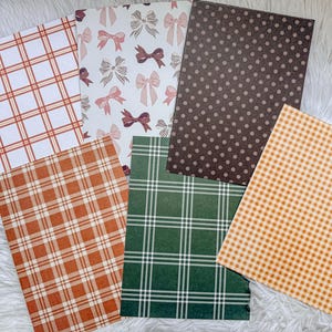 Op de afbeelding: Een verzameling papieren met patronen. Ontwerpen zijn onder meer oranje en rode ruitjes, roze en bordeauxrode strikken, bruine stippen, groen-witte ruitjes en oranje gingham. De papieren zijn gerangschikt op een wit, pluizig oppervlak.