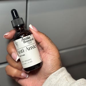 May include: A brown glass tincture bottle with a black dropper is held in a hand. The label reads "Avaia Botanicals Anti-Anxiety Tincture 100% Organic 59.15ml 2 oz."