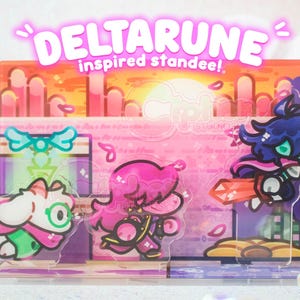 May include: A rectangular acrylic standee featuring a colorful sunset background and three cartoon characters. The text DELTARUNE inspired standee! is printed in white at the top. The characters include a white creature, a pink figure, and a dark-haired figure.