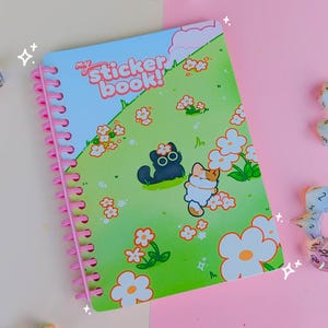 Puede incluir: Un libro de pegatinas reutilizable con una cubierta mate, que mide 12,7 cm x 17,8 cm. La portada presenta un campo verde con gatos y flores de dibujos animados. El libro tiene una encuadernación en espiral rosa. Los dados están esparcidos alrededor del libro.