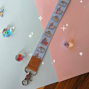 May include: A blue and white lanyard with a repeating pattern of cartoon creatures. The lanyard has a brown leather strap and a silver metal clip. The lanyard is on a pink and blue surface with dice.