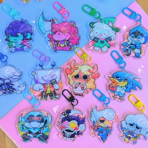 May include: A selection of colourful acrylic keyrings featuring cartoon characters in various designs. Each keyring has a metal clasp. The keyrings are arranged on a pink and blue surface.