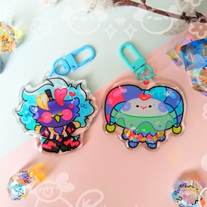 Puede incluir: Dos llaveros de personajes coloridos sobre un fondo azul y blanco. El llavero de la izquierda es un personaje azul, morado y amarillo con un corazón rojo y un sombrero de copa negro. El llavero de la derecha es un personaje verde, azul y morado con un gorro de bufón rojo y una cara blanca.