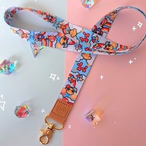 Op de afbeelding: Een blauwe en witte lanyard met een kleurrijk cartoonontwerp met ballonnen, regenbogen en lachende gezichten. De lanyard heeft een gouden metalen clip en een bruin leren bandje.