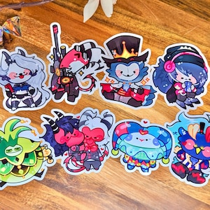 May include: A set of nine colorful cartoon character stickers. The stickers feature a variety of characters, including a green plant, a red and black demon, a blue and white jester, a blue and white bird, a white and gray wolf, a red and black dragon, a blue and white clown, a blue and white mermaid, and a blue and white robot.