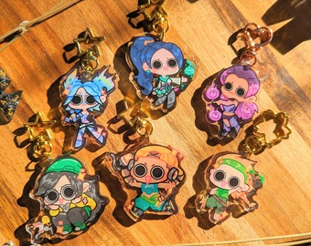 Può includere: Sei portachiavi con personaggi dei cartoni animati colorati con ciondoli a forma di stella dorati e anelli di catena dorati. I personaggi sono disegnati in stile chibi con occhi grandi e sono posizionati su uno sfondo di legno.