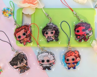 May include: Six colourful cartoon character phone charms with different designs. Each charm is attached to a string with a metal ring. The charms are displayed on a green phone case and a pink surface.