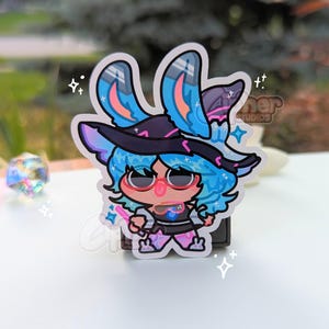 May include: A blue and pink cartoon character with bunny ears wearing a witch hat and holding a wand. The character is standing on a white background with sparkles.