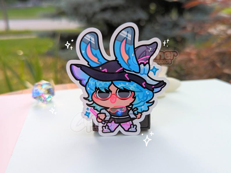 May include: A blue and pink cartoon character with bunny ears wearing a witch hat and holding a wand. The character is standing on a white background with sparkles.