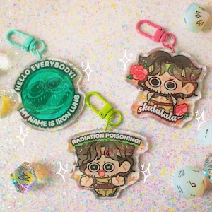 May include: Three acrylic keychains with colorful clasps and several multi-sided dice. One circular keychain is green with the text HELLO EVERYBODY! MY NAME IS IRON LUNG. Two character keychains feature text reading chalalala and RADIATION POISONING!