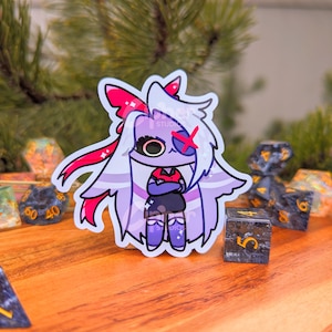 May include: A sticker of a cartoon character with white hair, a red bow, and a red X over one eye. The character is wearing a black dress and has their arms crossed. The sticker is on a wooden surface with several dice.
