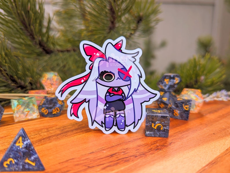 May include: A sticker of a cartoon character with white hair, a red bow, and a red X over one eye. The character is wearing a black dress and has their arms crossed. The sticker is on a wooden surface with several dice.