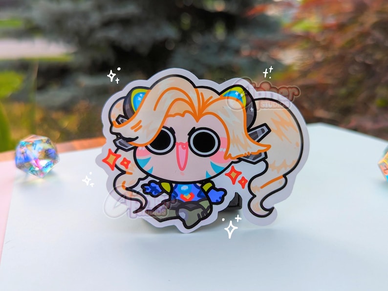 May include: A sticker of a cartoon character with long blonde hair, wearing a blue and yellow outfit. The character has large eyes and a wide smile. The sticker is on a white background with sparkles.